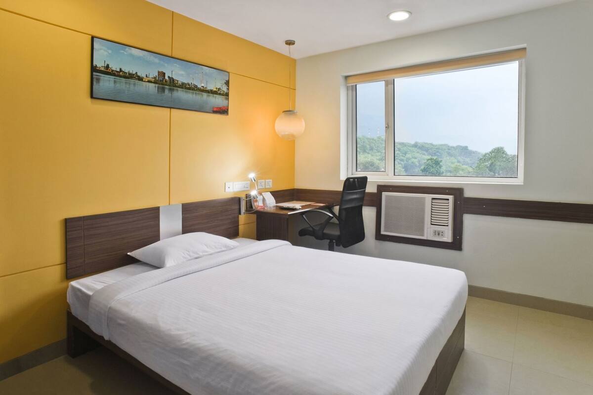 A hotel room with a bed, a desk with a chair, a window with a view, and a wall-mounted air conditioner.