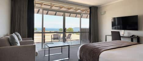 A hotel room with a bed, a desk, a chair, a sofa, and a large window with a view of the sea.