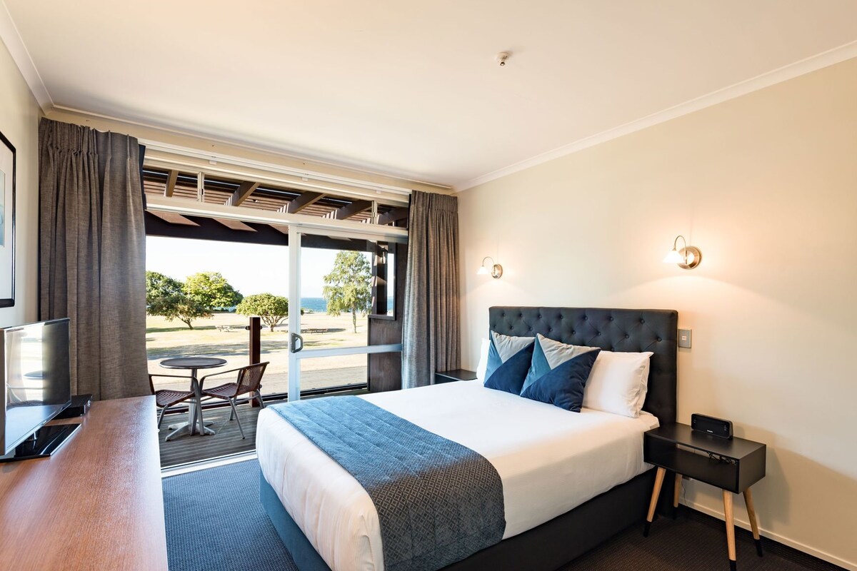 A hotel room with a bed, a desk, a chair, a TV, and a view of the outdoors.