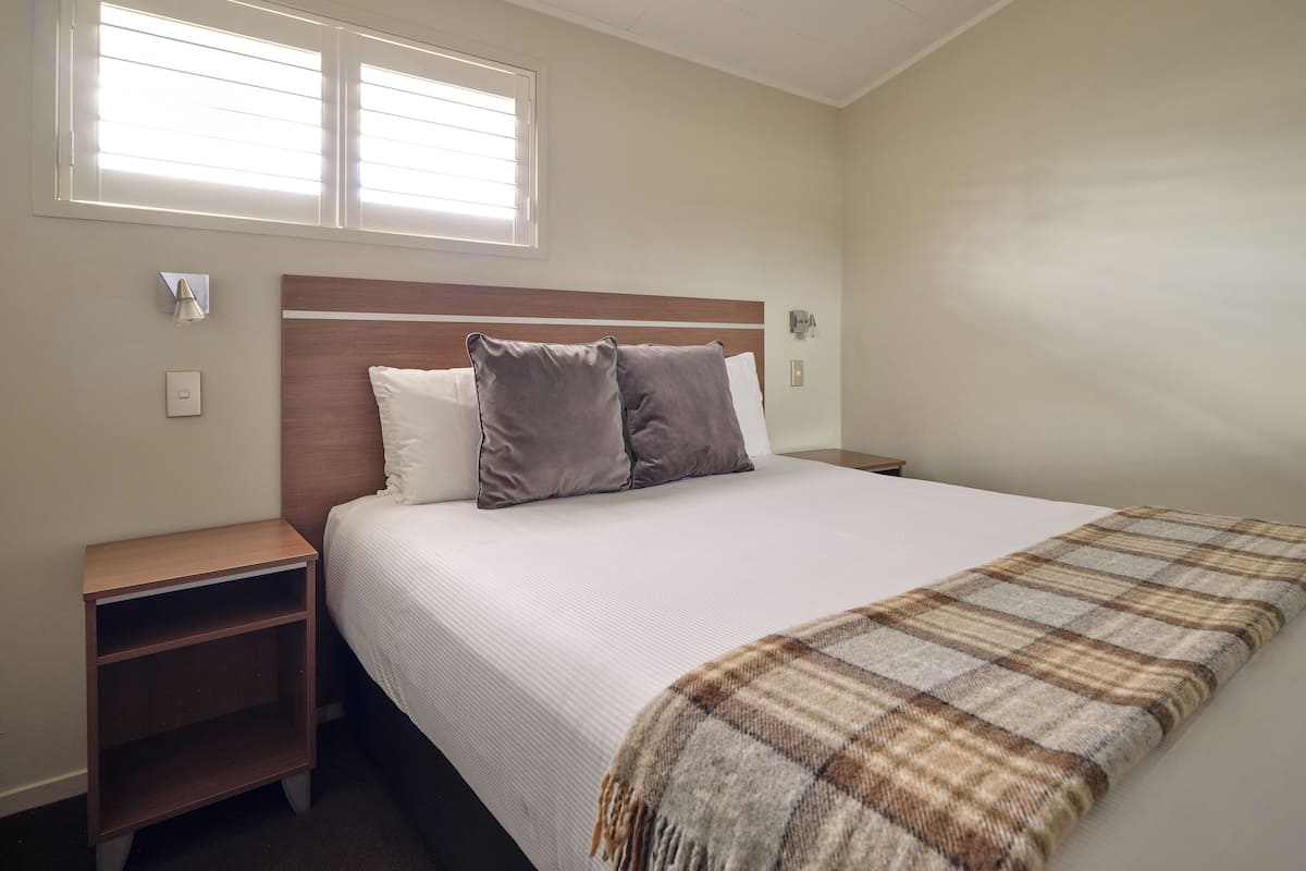 Een slaapkamer met een bed, twee nachtkastjes, een venster met zonwering en een wandlamp.