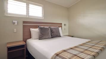 A bedroom with a bed, two bedside tables, a nightstand, a window with blinds, and a wall-mounted lamp.