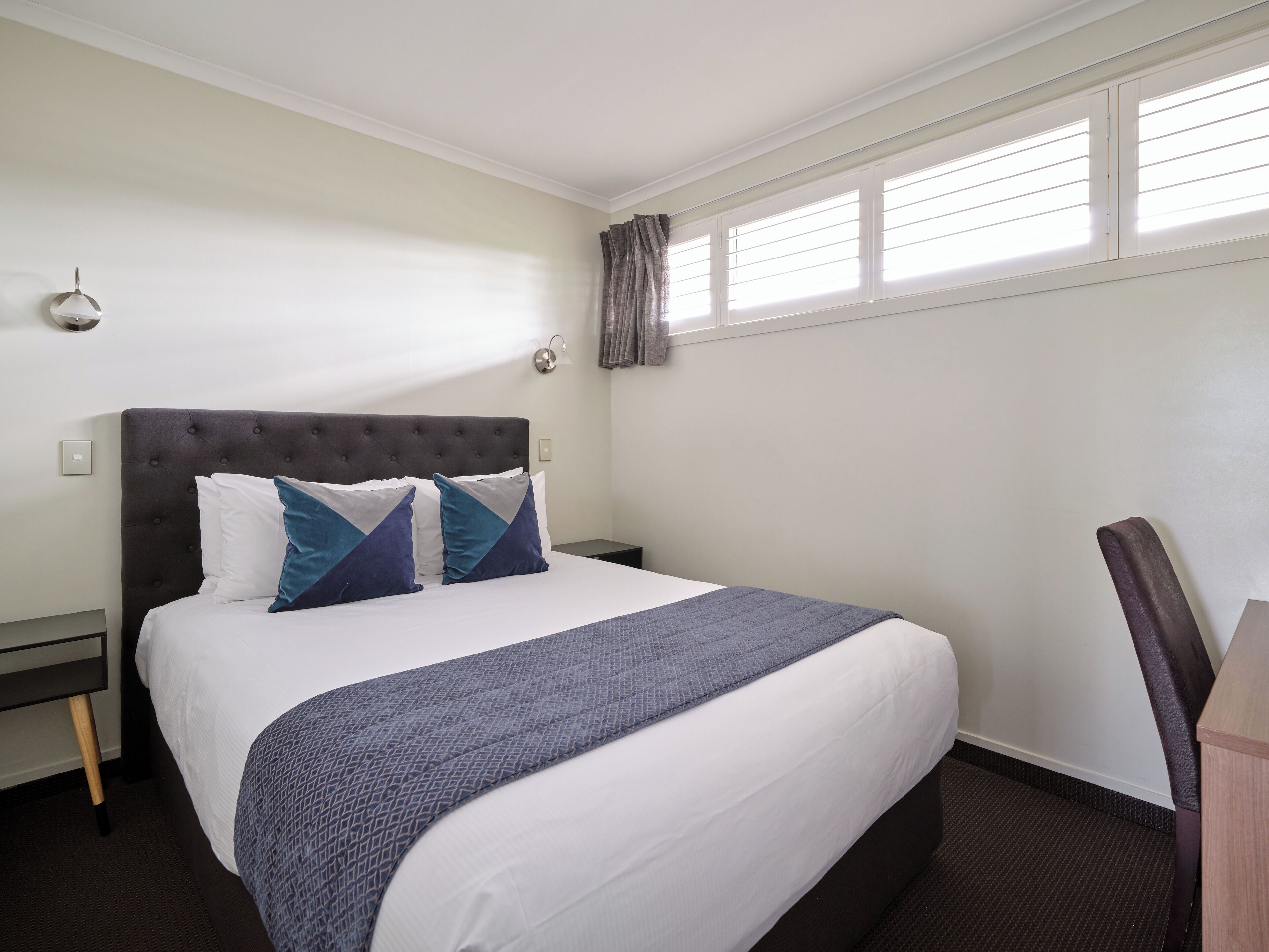 A neatly made bed with a dark headboard, two pillows, and a blue blanket. There is a nightstand with a lamp, a window with blinds, and a chair.