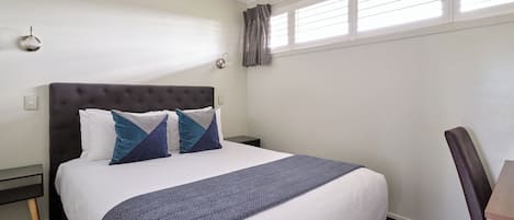 A neatly made bed with a dark headboard, two pillows, and a blue blanket. There is a nightstand with a lamp, a window with blinds, and a chair.