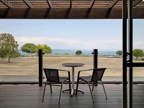 A covered outdoor seating area with two chairs and a table, overlooking a beach with parked cars and a clear view of the ocean. - Suncourt Hotel & Conference Centre (Taupo)