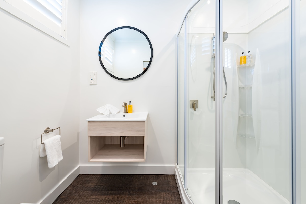 Une salle de bain moderne avec une douche à parois de verre, un miroir rond et un lavabo blanc avec un meuble-vasque en bois.