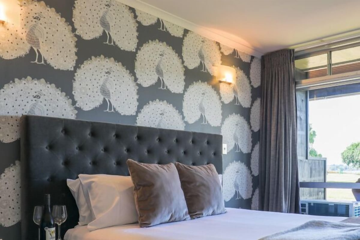 A hotel room with a tufted headboard, a bed with white linens and two pillows, a bedside table with a wine bottle and glasses, and a window with curtains.
