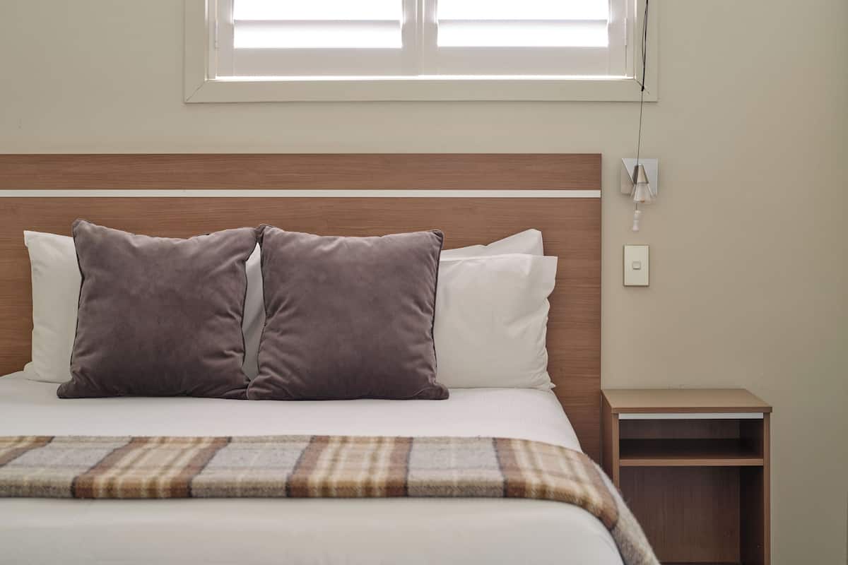 Een net opgemaakt bed met twee kussens, een nachtkastje en een raam met zonwering.