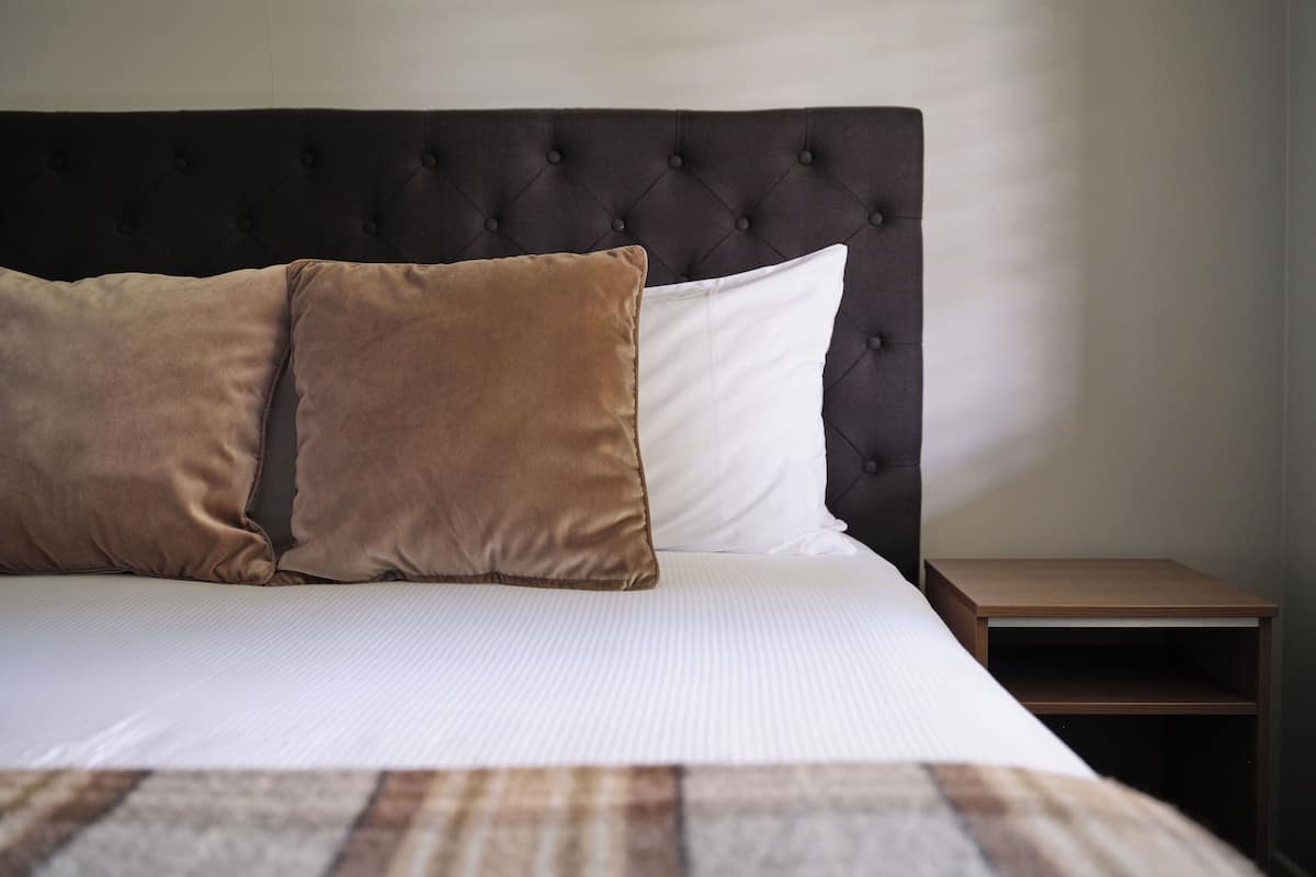 Een net opgemaakt bed met een gestuufd hoofdbord, twee kussens en een houten nachtkastje ernaast.