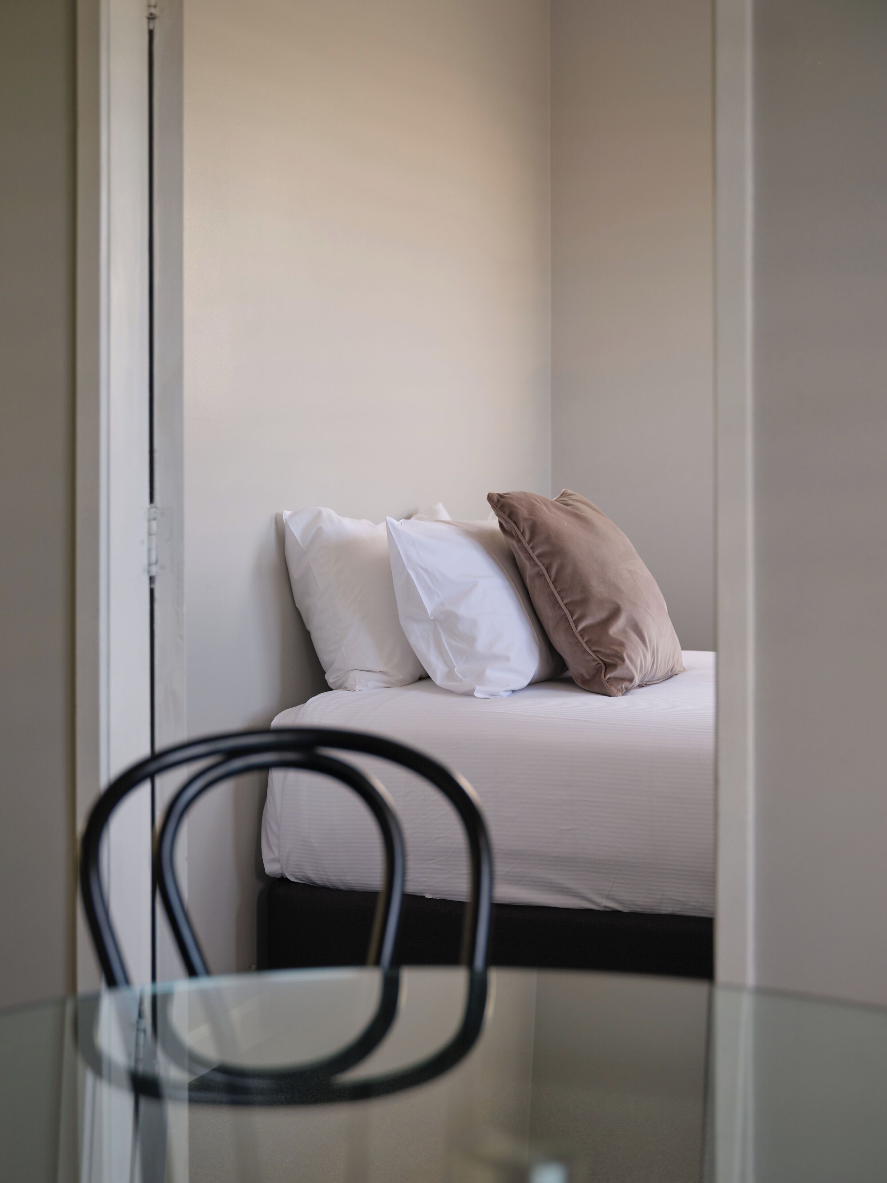 Un lit avec des oreillers blancs et beige, une table en verre avec une chaise, et un mur blanc.