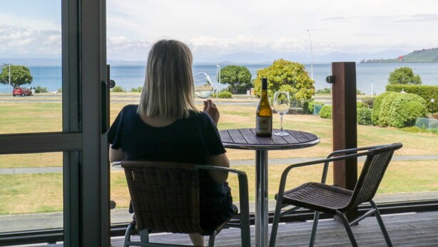 Una persona sentada a una mesa con una botella de vino y una copa, con vistas a un paisaje pintoresco de un lago y colinas.