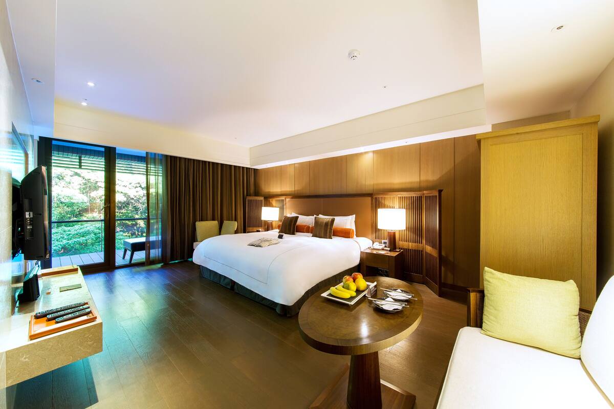 Quarto de hotel com uma cama grande, mesas de cabeceira, uma escrivaninha com cadeira, uma mesa redonda com frutas e vista para o verde pela janela.