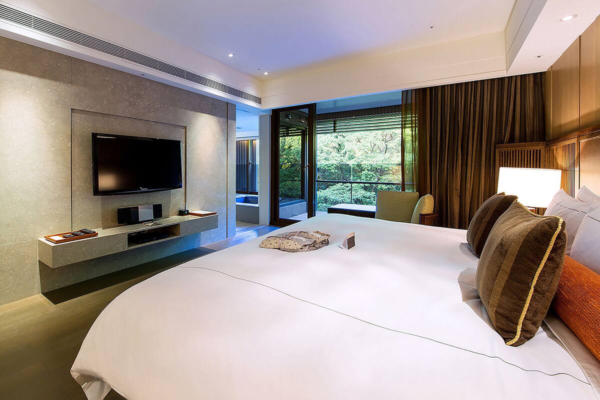 Quarto de hotel moderno com uma cama grande, televisão de tela plana na parede, área de estar com vista para o exterior e uma janela ampla com cortinas.
