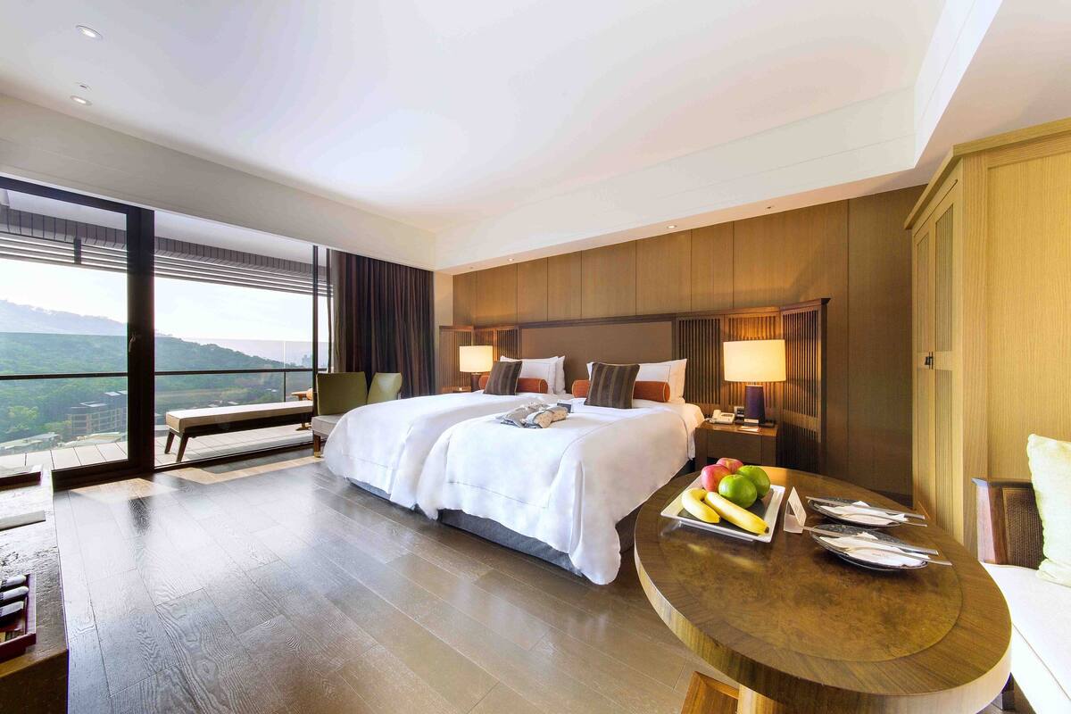 Quarto de hotel com uma cama grande, uma mesa de jantar com frutas e vista para o exterior.