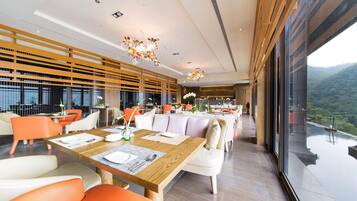 A modern dining area with wooden tables, orange and white chairs, and large windows offering a view of the mountains.