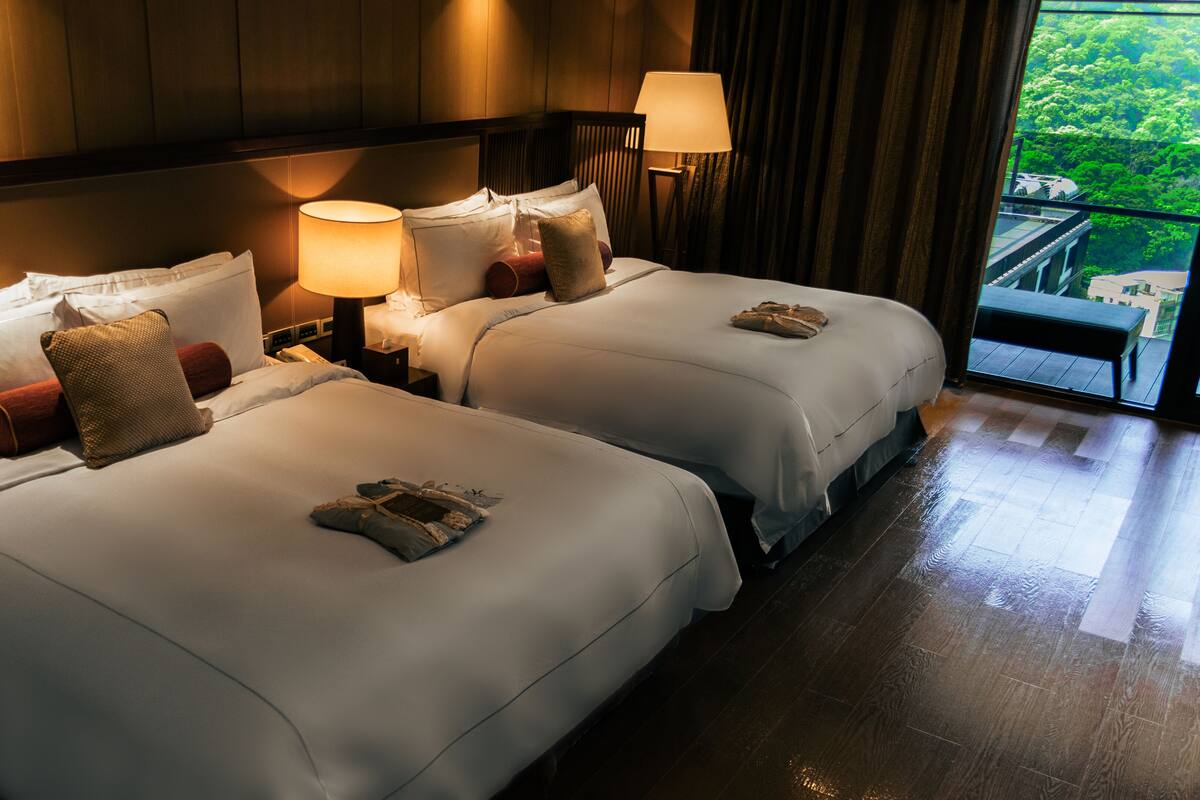 Quarto de hotel com duas camas, uma mesa de cabeceira com abajur e vista para uma varanda com vegetação.