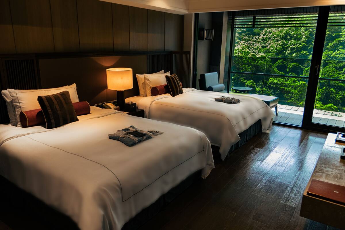 Quarto de hotel com duas camas, mesa de cabeceira, abajur, mesinha e vista para o verde através de amplas janelas.