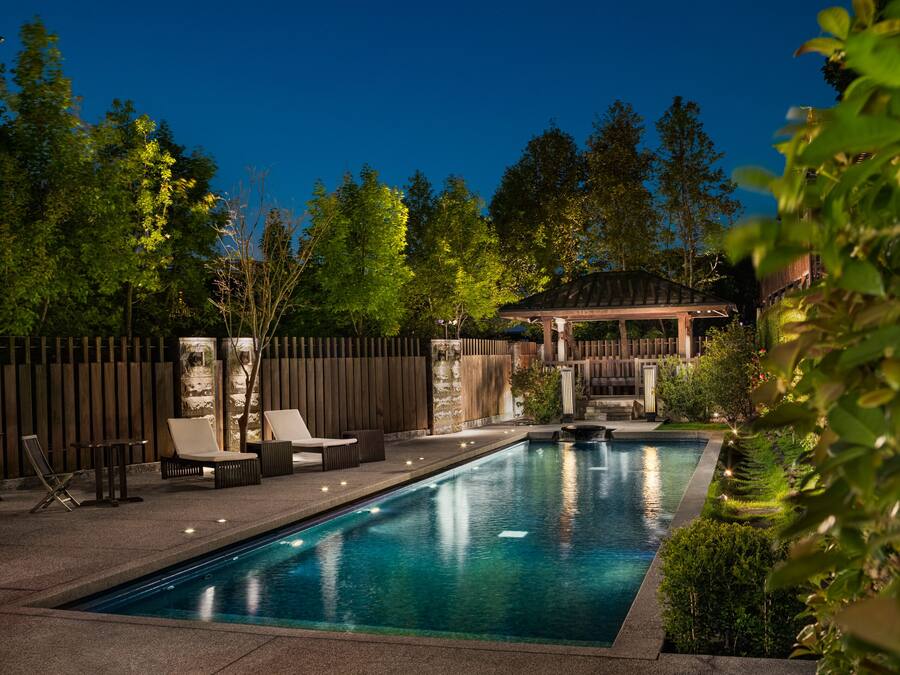 Une piscine rectangulaire dotée d’une terrasse en bois et de chaises longues, entourée d’une clôture en bois et d’arbres.