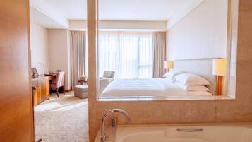 Une chambre d’hôtel avec un grand lit, un bureau, une chaise, un canapé, une salle de bain avec une baignoire et un miroir qui reflète la pièce.