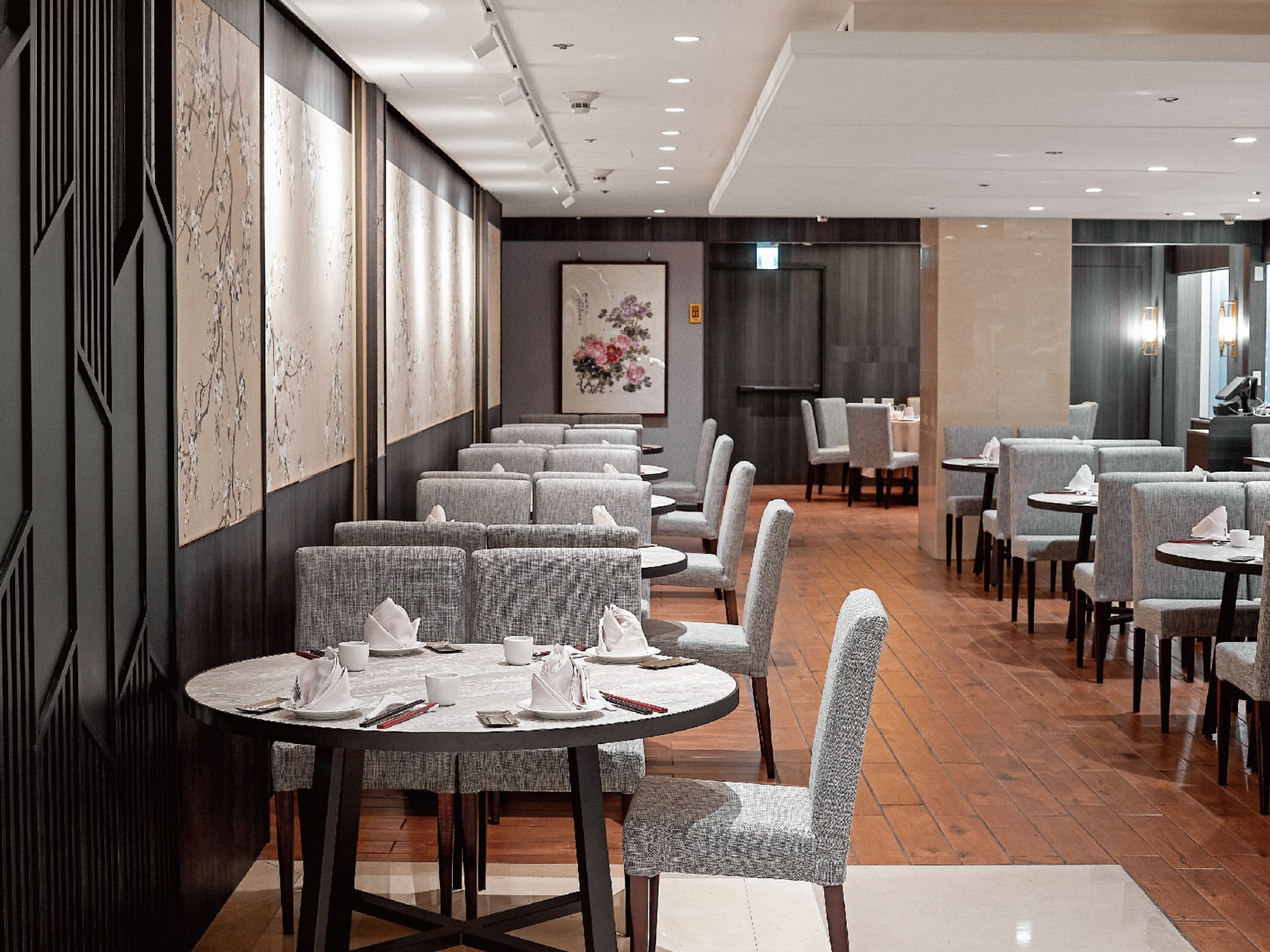 A modern restaurant with round tables and grey upholstered chairs, featuring a wooden floor and a large wall art piece.