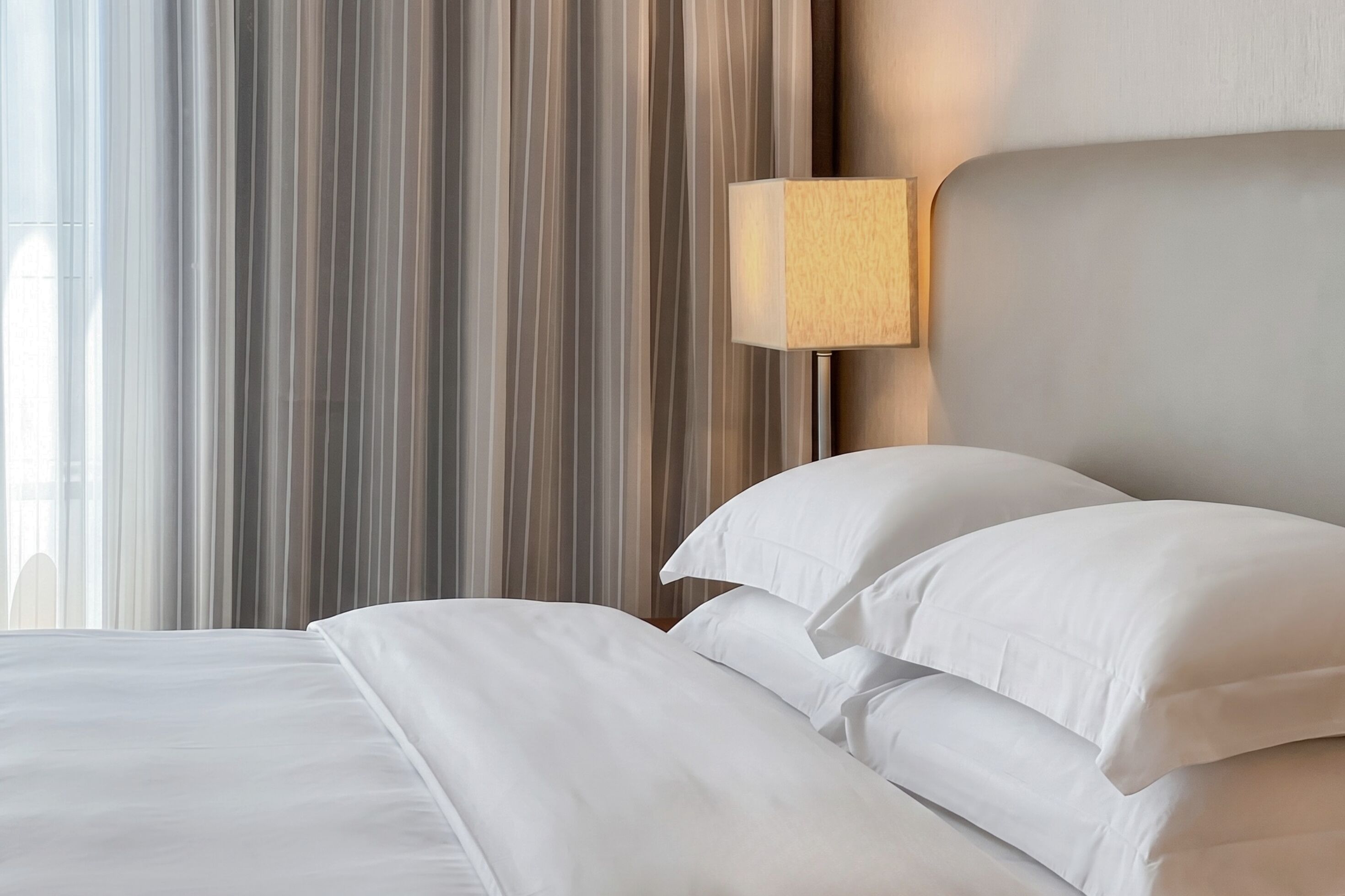 A neatly made bed with white linens and pillows, a wooden headboard, and a wall-mounted light fixture.