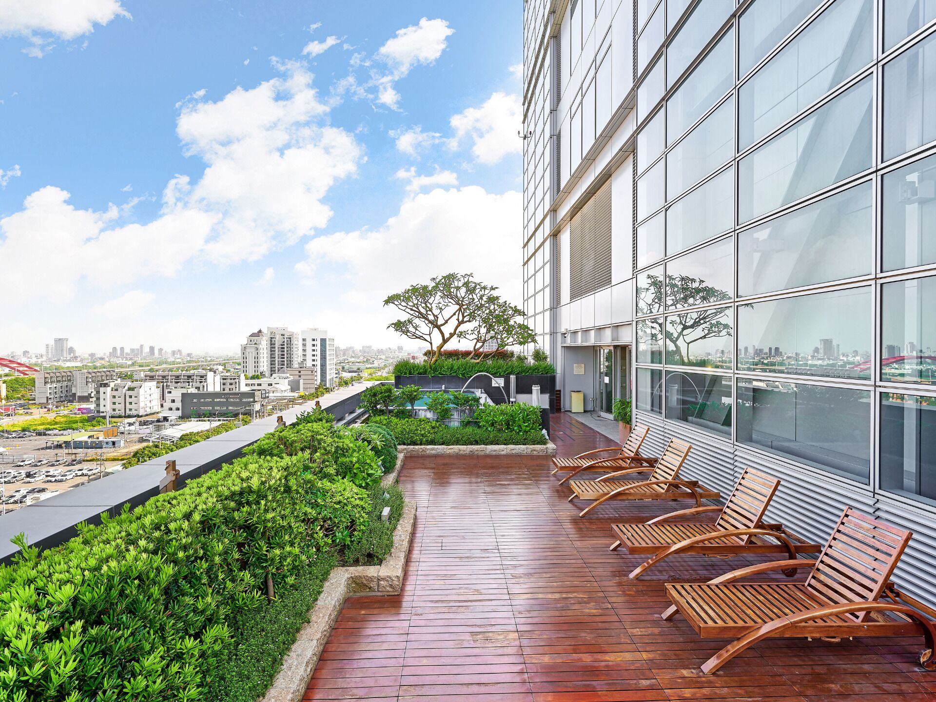 A rooftop terrace with a row of wooden lounge chairs, a small garden area, and a cityscape view.