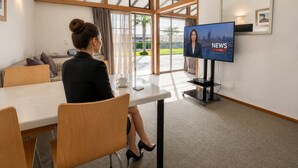 A woman sitting at a table with a television displaying 'NEWS LIVE' in a modern room with large windows and a view of greenery outside. - Teal Motor Lodge (Gisborne)