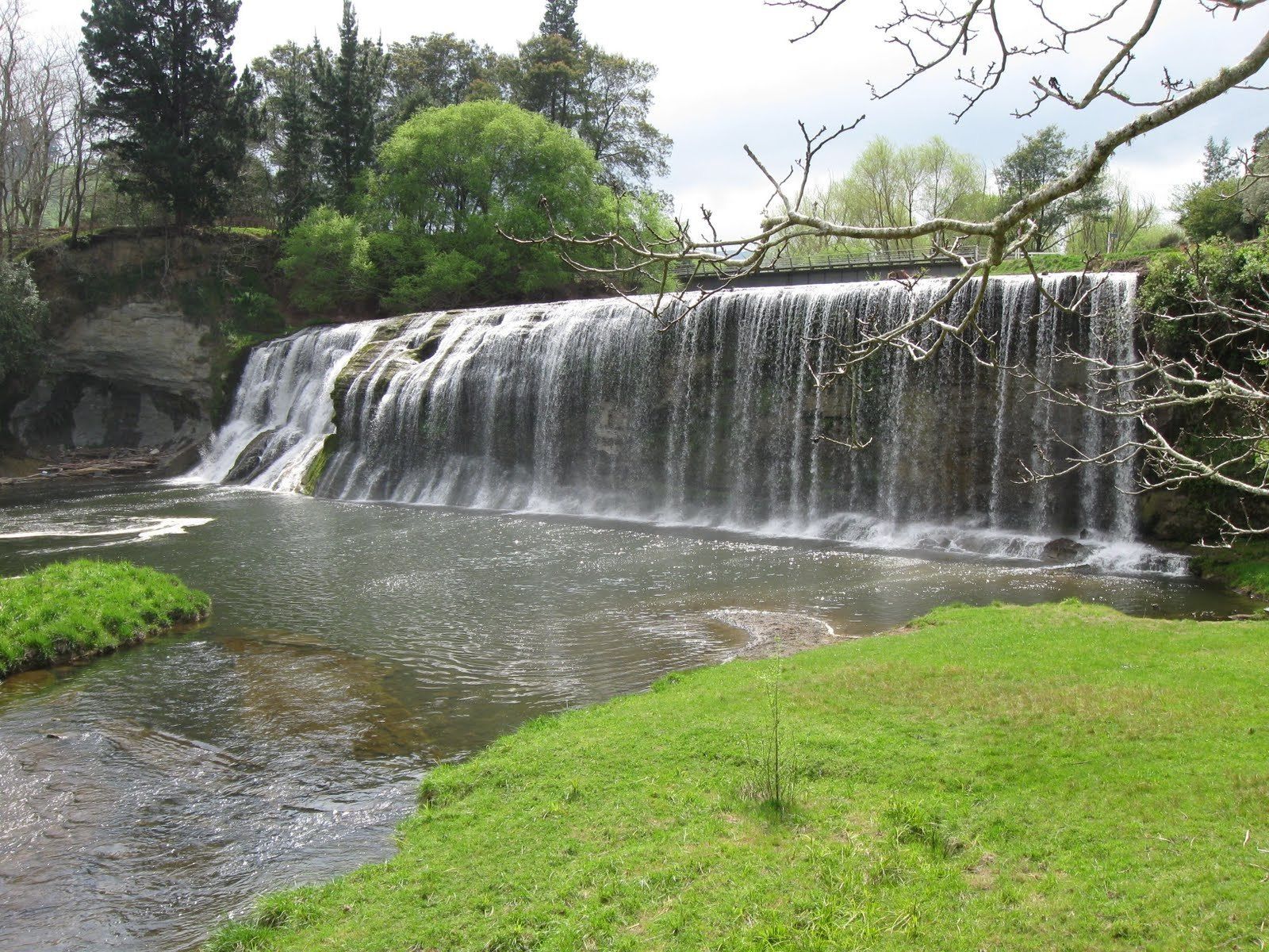 A waterfall cascading over a rocky ledge into a river, surrounded by lush greenery and trees.