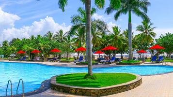 A swimming pool with lounge chairs and red umbrellas, surrounded by palm trees.