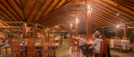 An indoor dining area with wooden tables and chairs, under a high wooden ceiling with exposed beams.