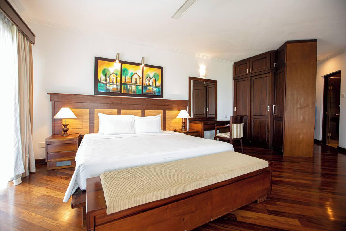 Una habitación de hotel con una cama grande, cabecera de madera, dos mesitas de noche con lámparas, un aparador y un cuadro sobre la cama.