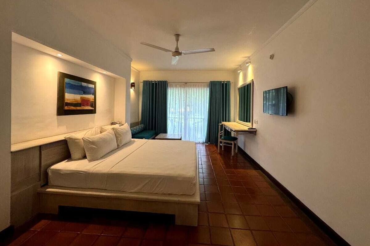 Habitación de hotel con cama, escritorio, silla, ventilador de techo y un cuadro en la pared.