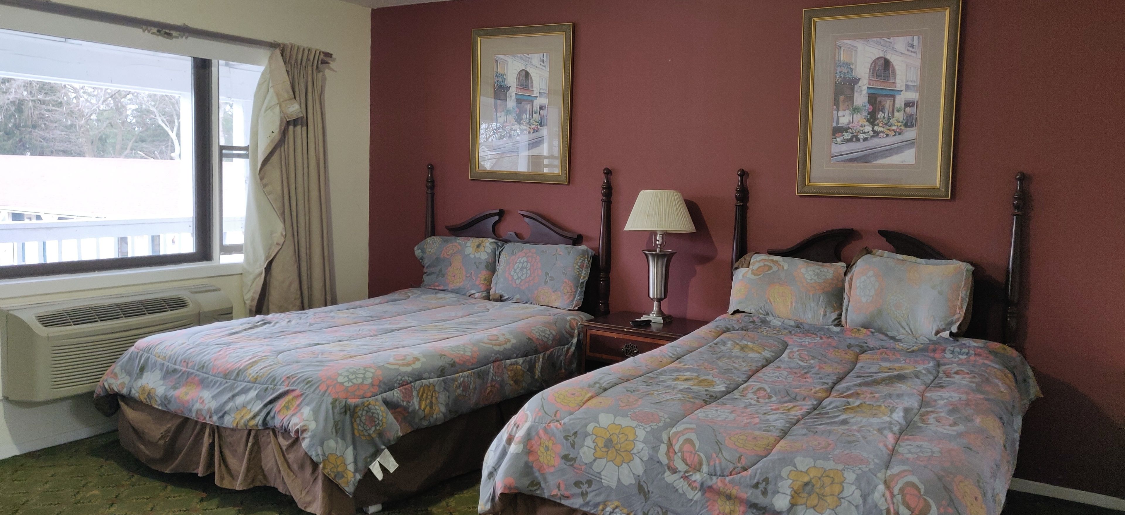 A hotel room with two beds, a window with curtains, a radiator, and two framed pictures on the wall.