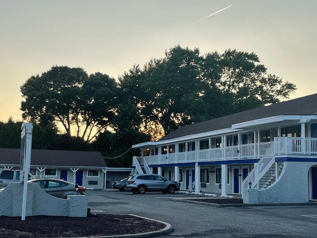 A motel with a two-story structure, a covered parking area, and a large tree in the background.