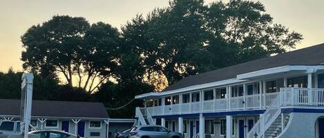 A motel with a two-story structure, a covered parking area, and a large tree in the background.