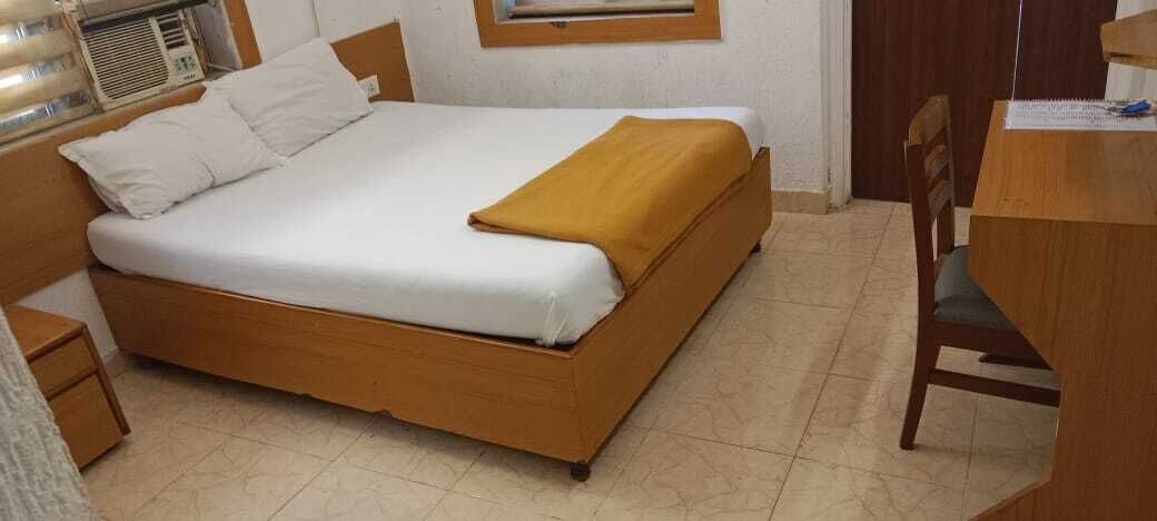 Una cama individual con estructura de madera y ropa de cama blanca, una mesita de noche pequeña y una ventana con persianas.