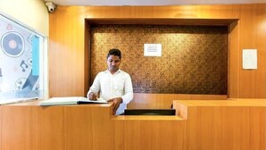 A reception desk with a wooden finish, a person seated behind it, and a wall with a textured pattern. - Hotel Red Palm (Mumbai)