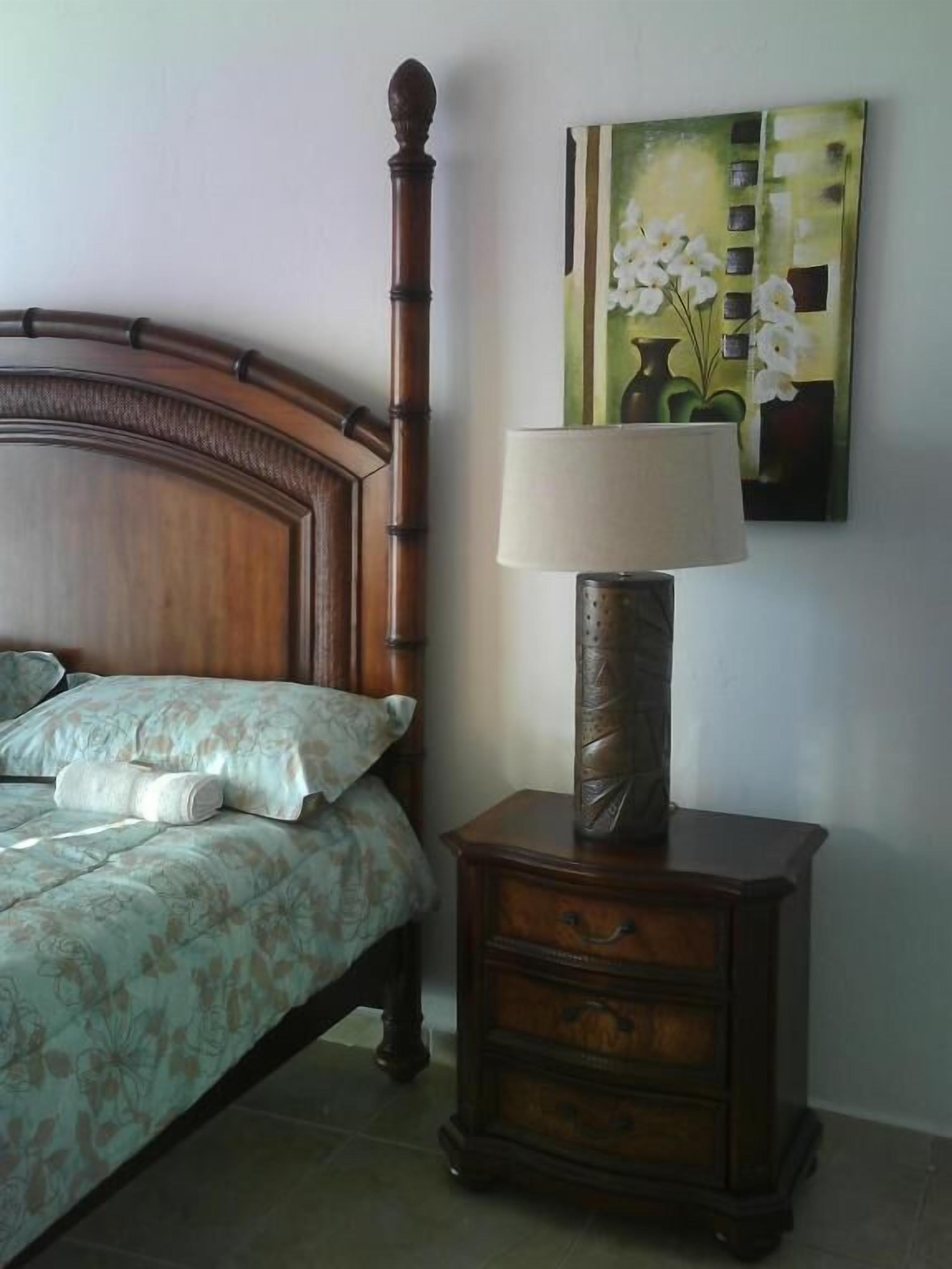 A neatly made bed with floral bedding, a wooden nightstand with a lamp, and a painting of flowers on the wall.