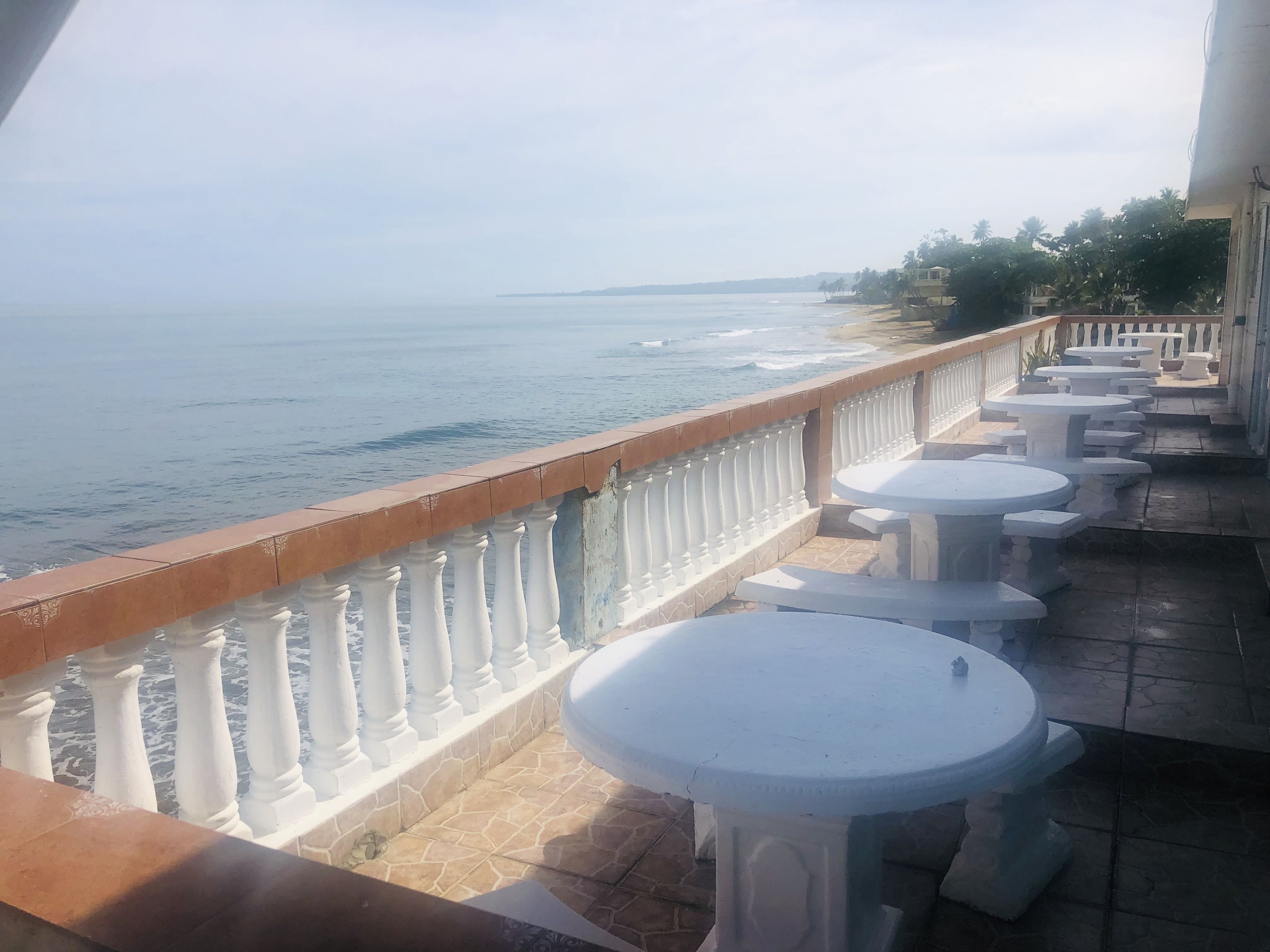 Un balcón con mesas blancas y barandilla, con vista al mar.