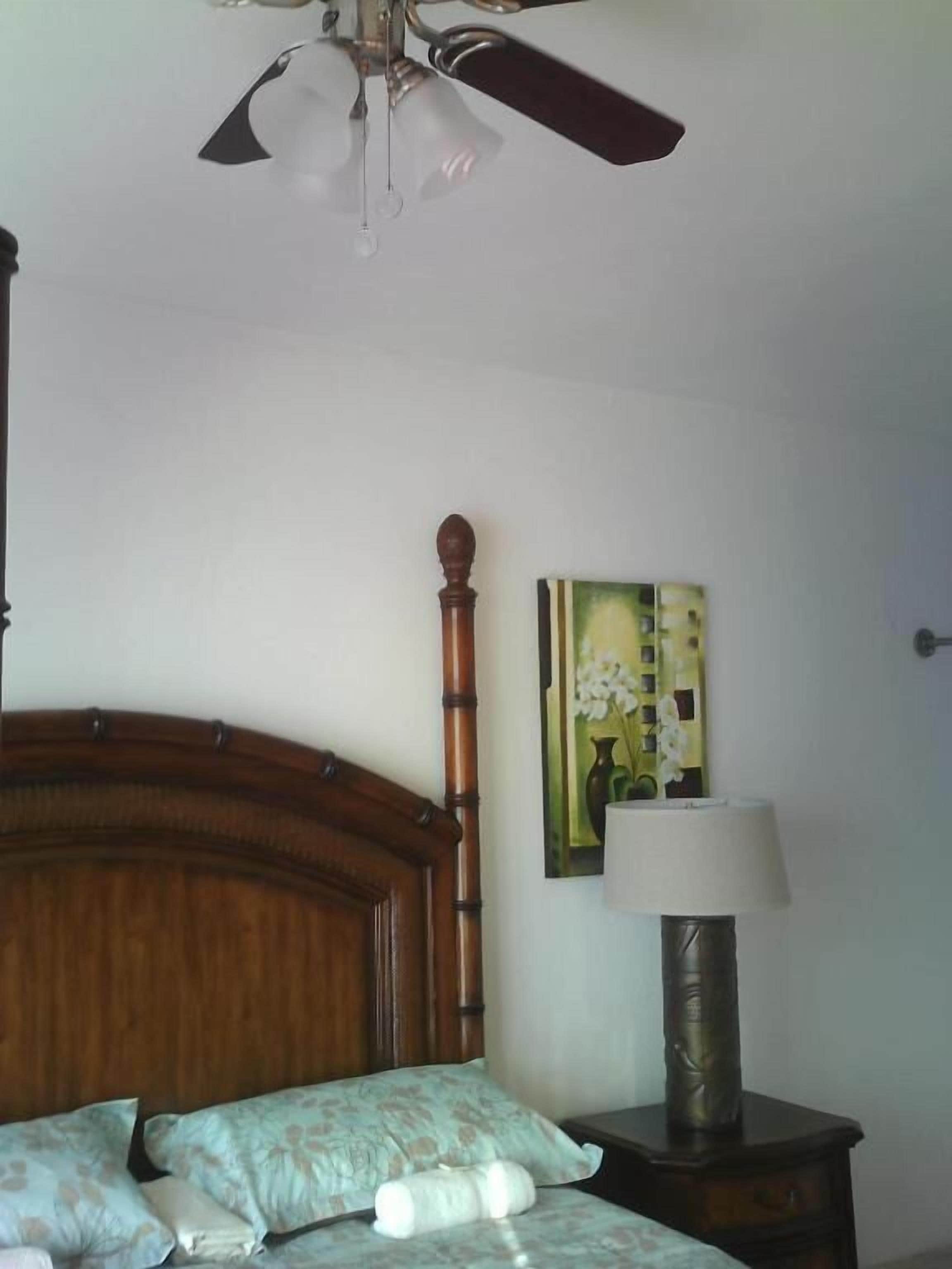 Un dormitorio con cabecera de madera, cama con ropa de cama floral, mesita de noche con lámpara y ventilador de techo.