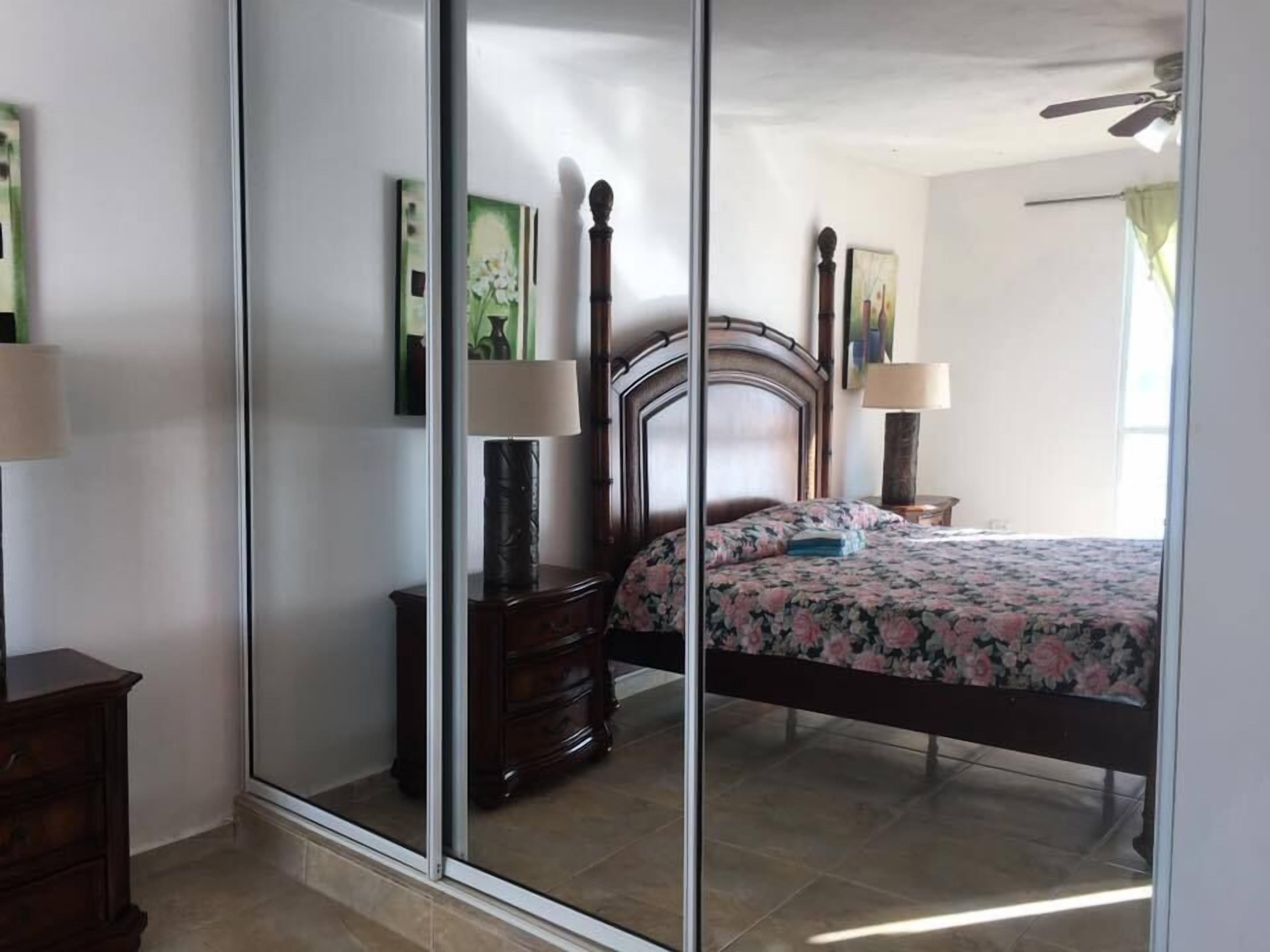 A bedroom with a large mirror, a wooden bed frame, a floral bedspread, and a nightstand with a lamp.