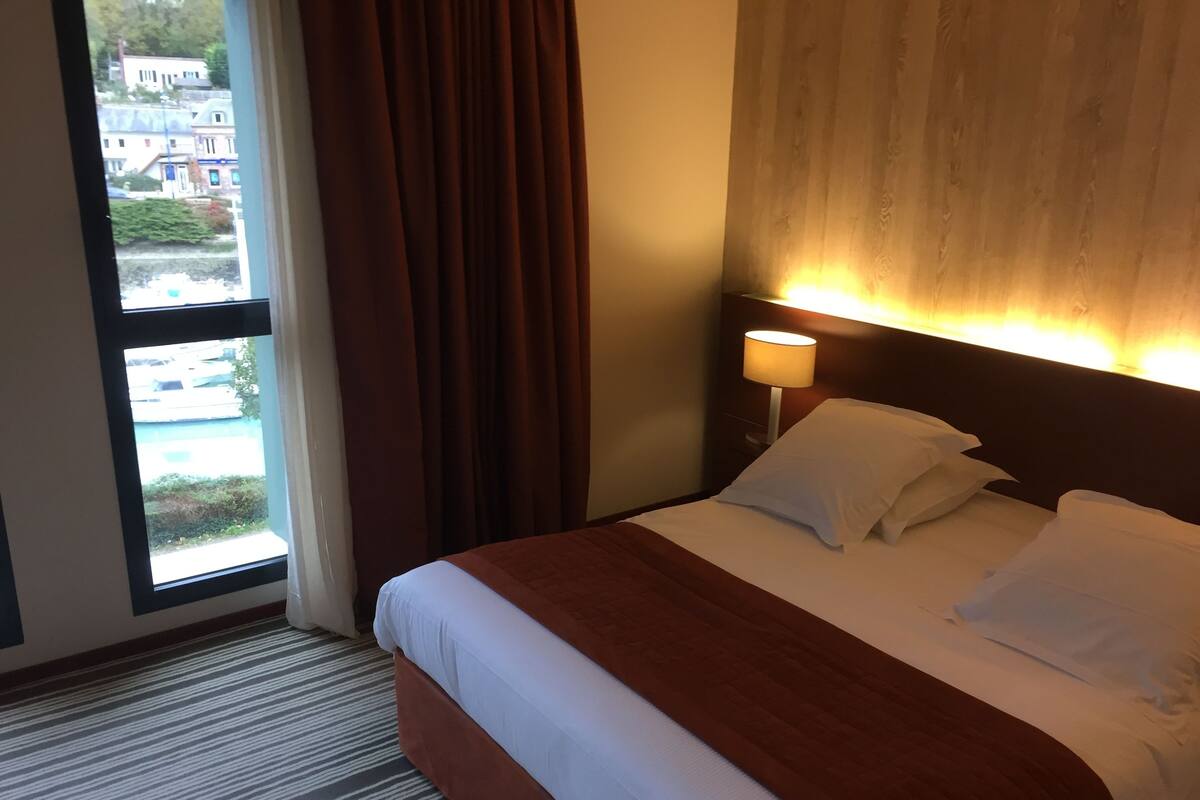 A hotel room with a bed, a nightstand with a lamp, a window with curtains, and a view of buildings outside.
