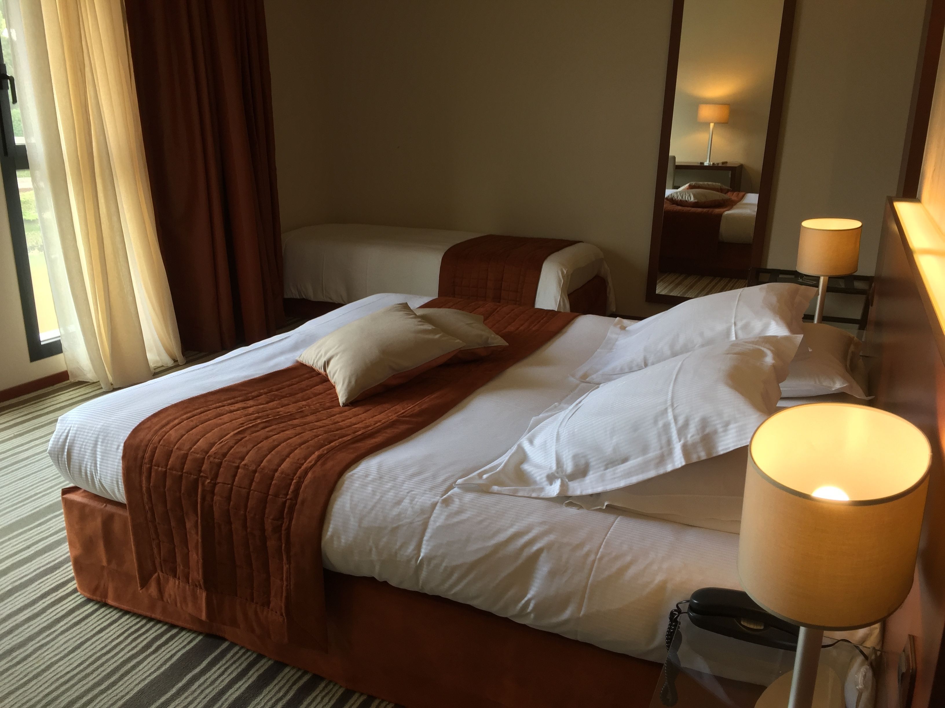 Habitación de hotel con una cama grande, una cama más pequeña, una mesita de noche con lámpara y un espejo en la pared.