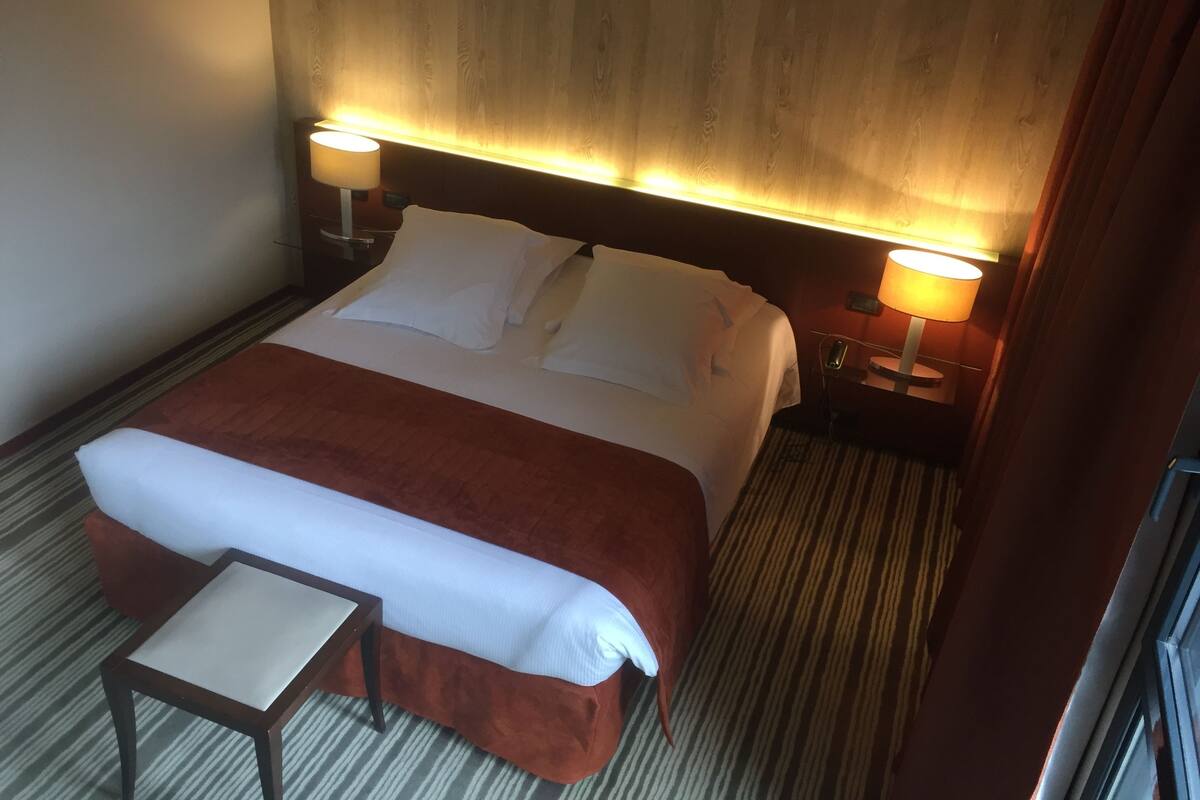 A hotel room with a bed, two bedside lamps, a small table, and a window.