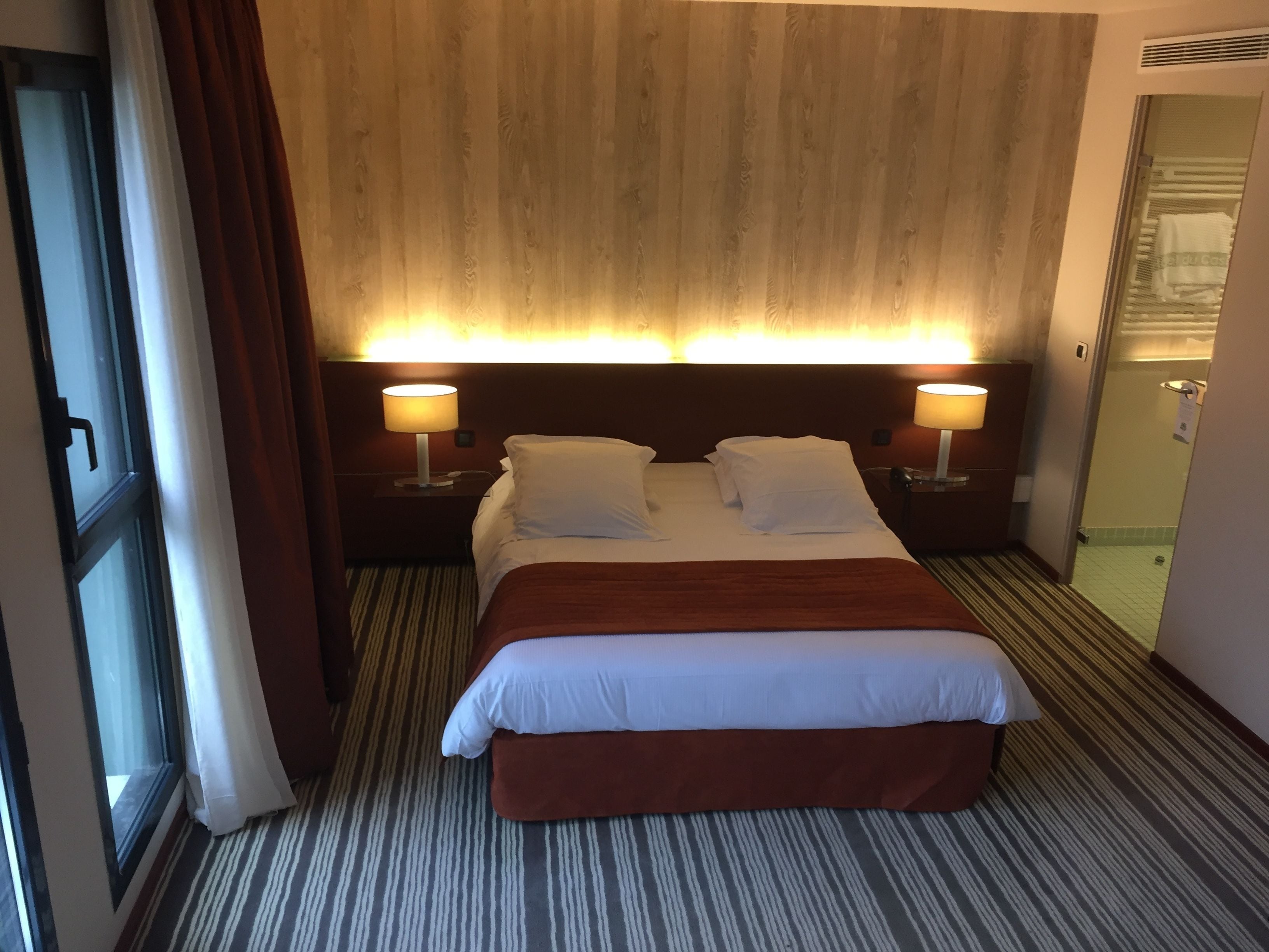 Une chambre d’hôtel avec un grand lit, deux lampes de chevet, une tête de lit en bois et une moquette à rayures.
