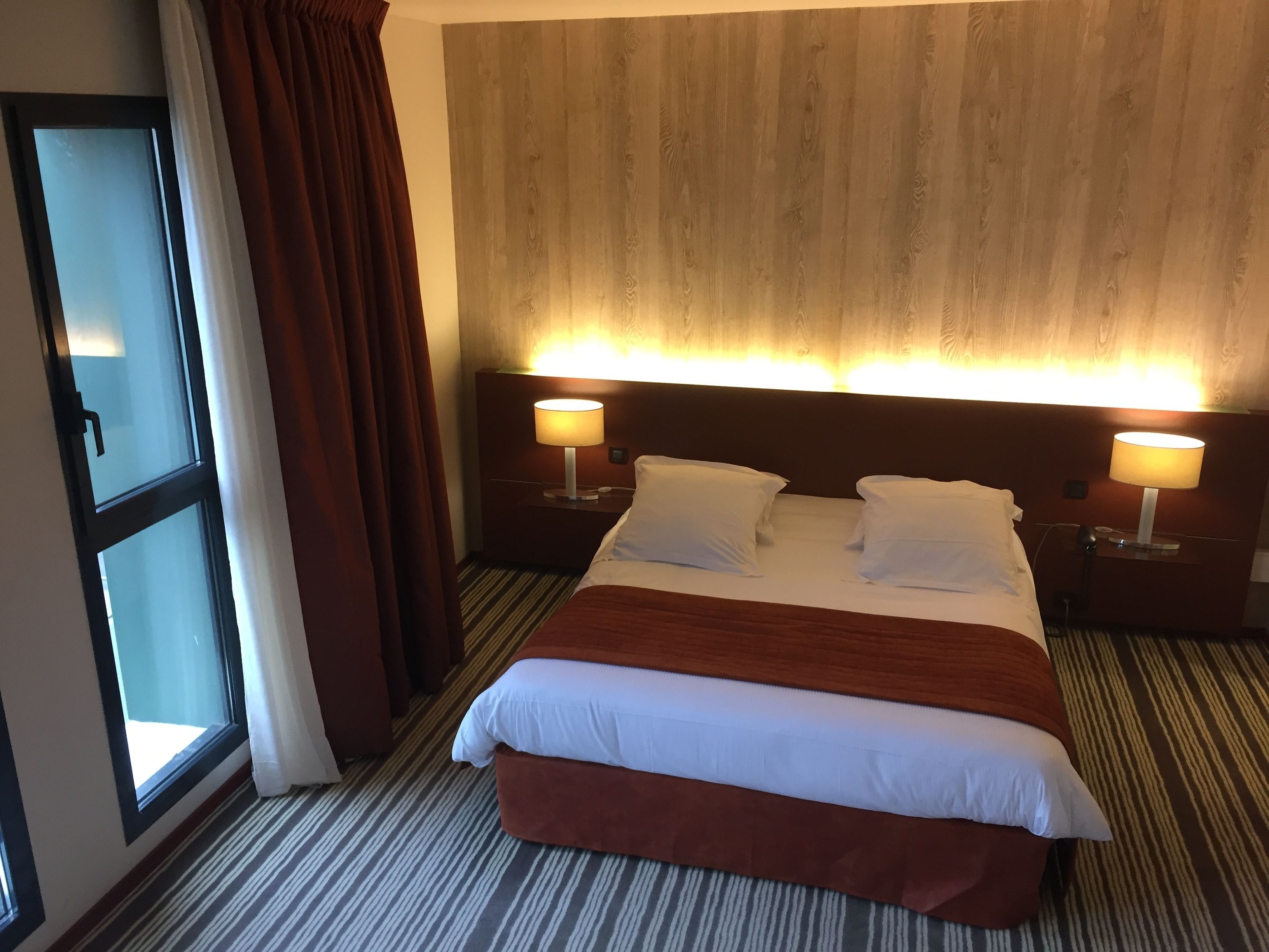 A hotel room with a large bed, two bedside tables with lamps, a window with curtains, and a wooden headboard.