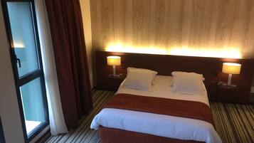 A hotel room with a large bed, two bedside tables with lamps, a window with curtains, and a wooden headboard.