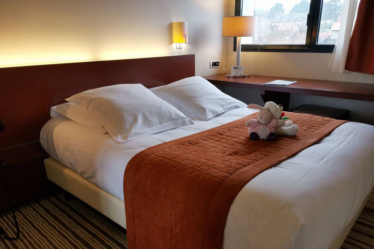 A hotel room with a bed, pillows, a blanket, a lamp, and a window.