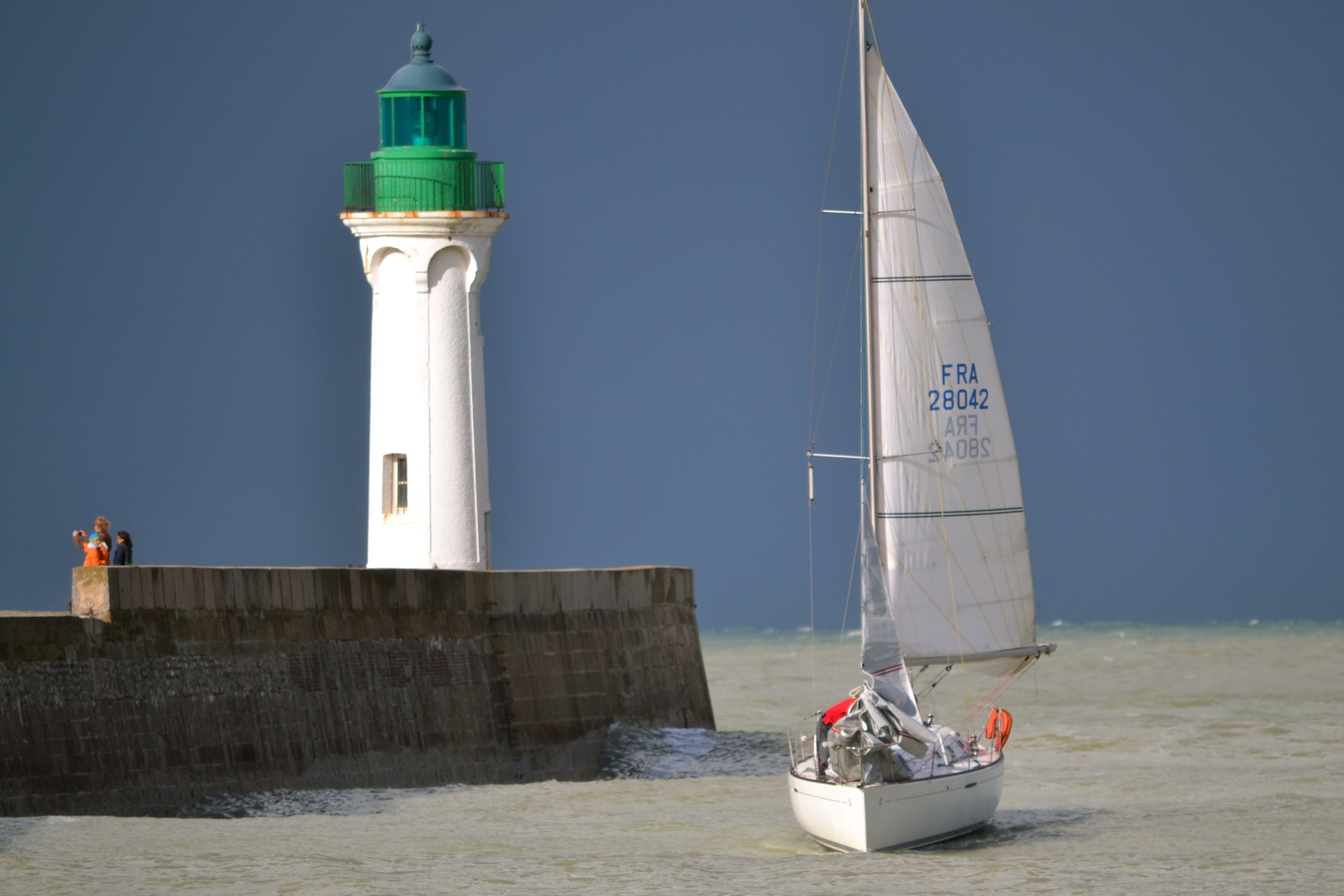 Un velero con casco blanco y toldo verde, navegando cerca de un faro con techo verde y base blanca, y una barrera de hormigón.