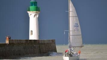 Un voilier à la coque blanche et au pont vert navigue près d’un phare également doté d’un pont vert et d’une base blanche, ainsi que d’une barrière en béton.