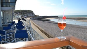 Ein Balkon mit Weinglas und Halm, Blick auf einen Strand mit Stühlen und Geländer.
